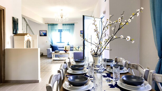 A dining area with a table set for four, a vase with flowers, and a view into a living room with a sofa and a TV.