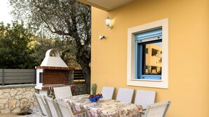 An outdoor dining area with a table set for four, surrounded by chairs, a decorative tablecloth, and a fruit bowl.