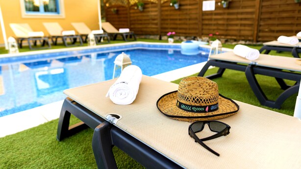 Un espace aménagé au bord de la piscine, avec des chaises longues, un parasol et un chapeau sur lequel est écrit « Grèce Rhodes ».
