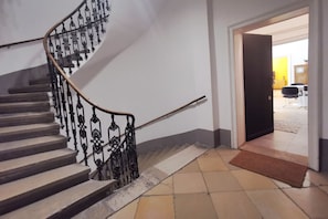 Un escalier orné de rampes noires menant à une pièce dont le sol est recouvert de carreaux, et une porte donnant sur une autre pièce.
