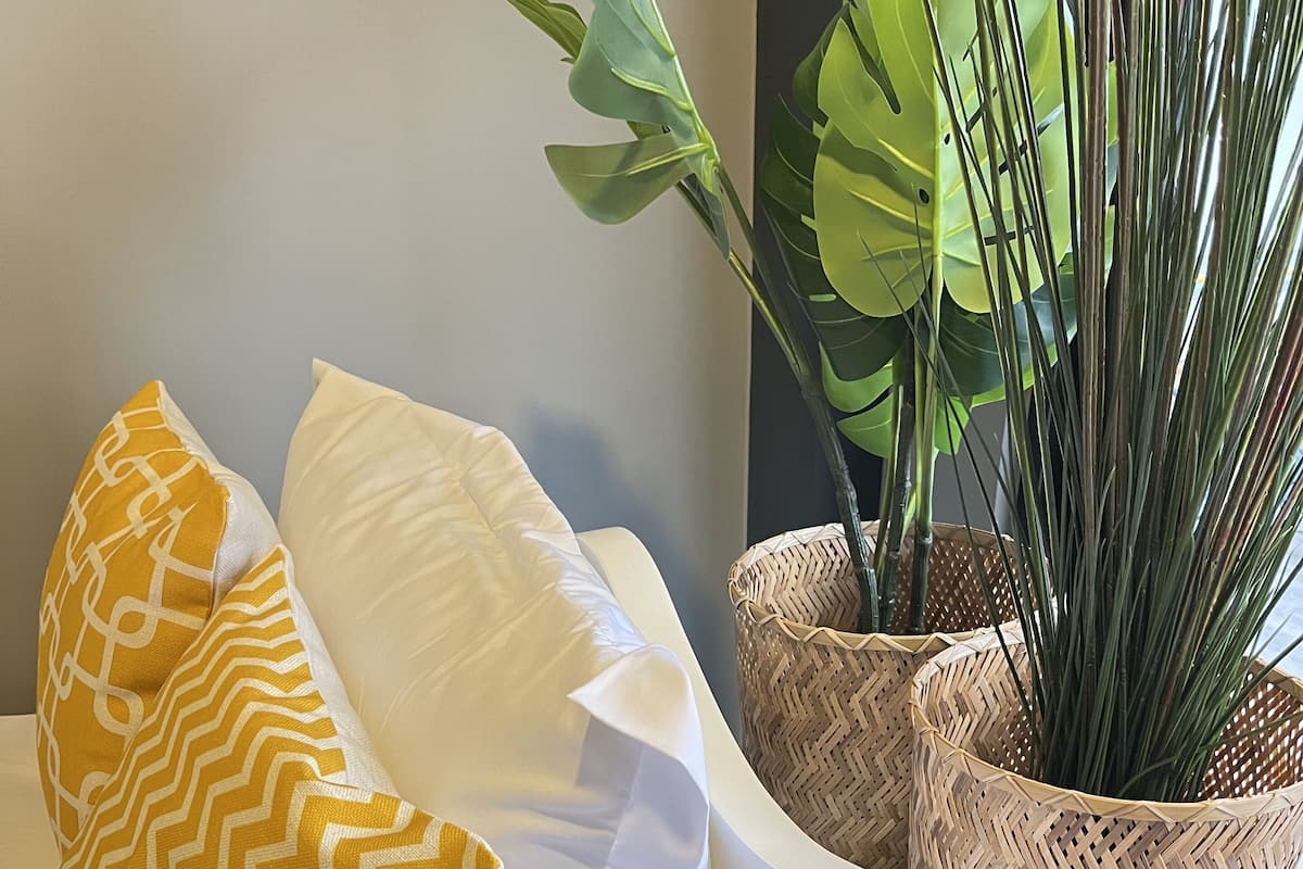 A white sofa with two pillows, one yellow and one white, and a potted plant in a woven basket.