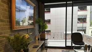 A balcony with a view of buildings, a wooden table, a chair, a potted plant, a framed picture, and a small wooden figure. - Hotel Puerta Roja Reforma  (Mexico City)