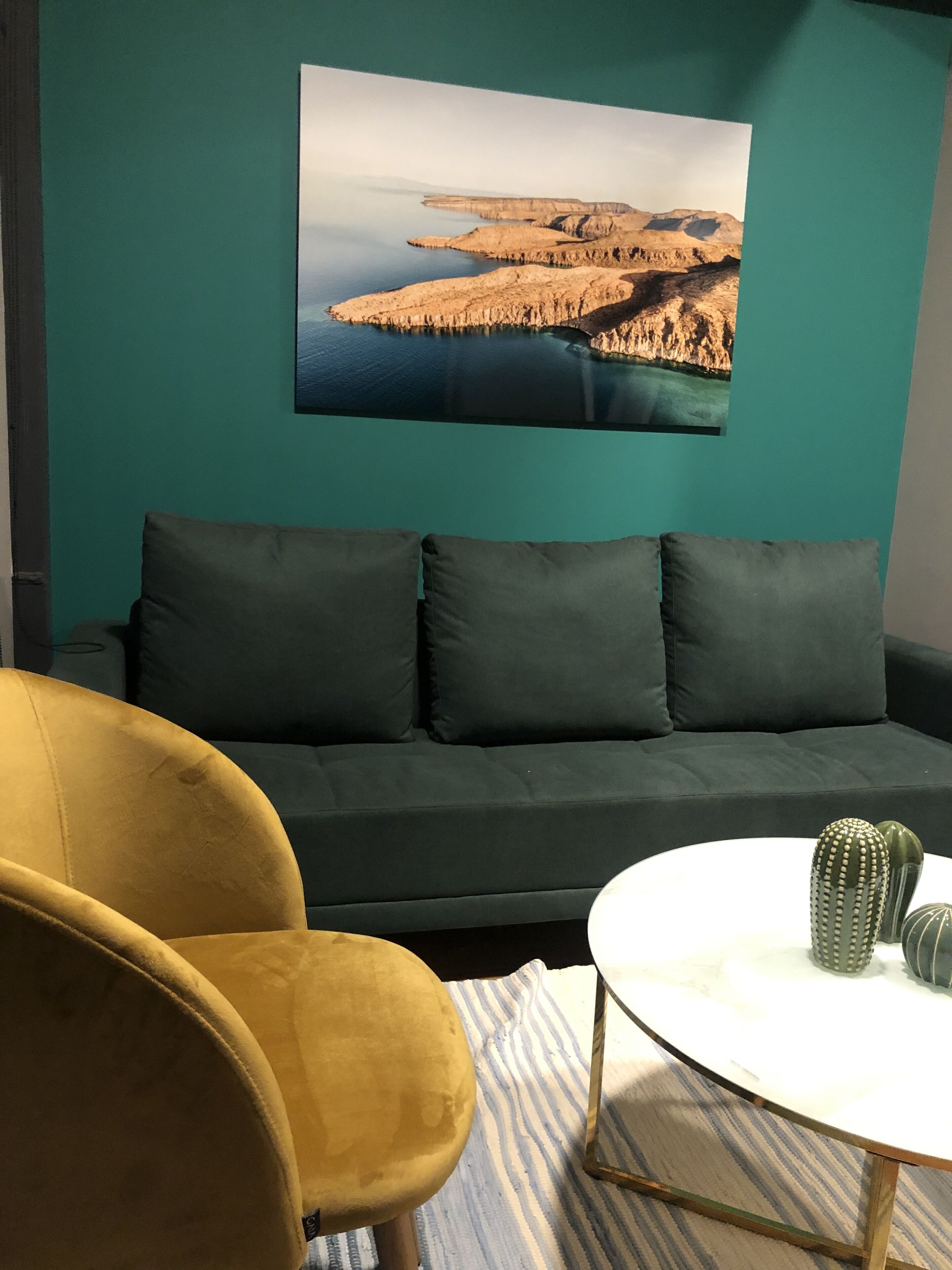 A teal-colored wall with a large landscape painting, a dark green sofa, a yellow armchair, and a small white table with two cacti.