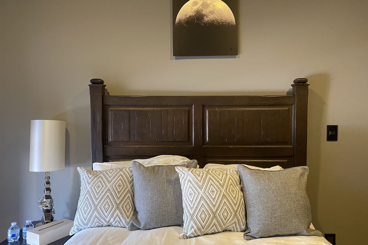 A neatly made bed with patterned pillows, a wooden headboard, a wall-mounted lamp, and a framed picture of the moon.