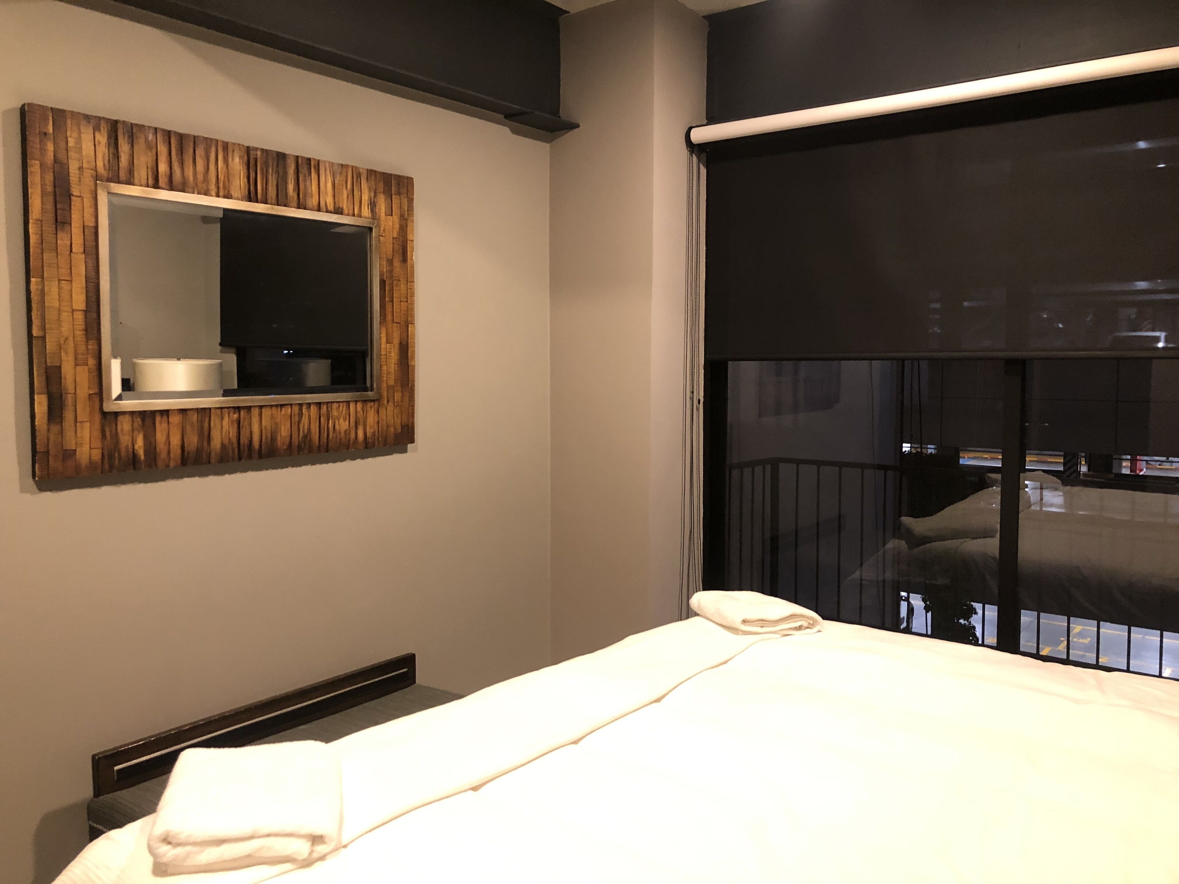 A hotel room with a bed, a wooden-framed mirror, and a window with a view of the cityscape.