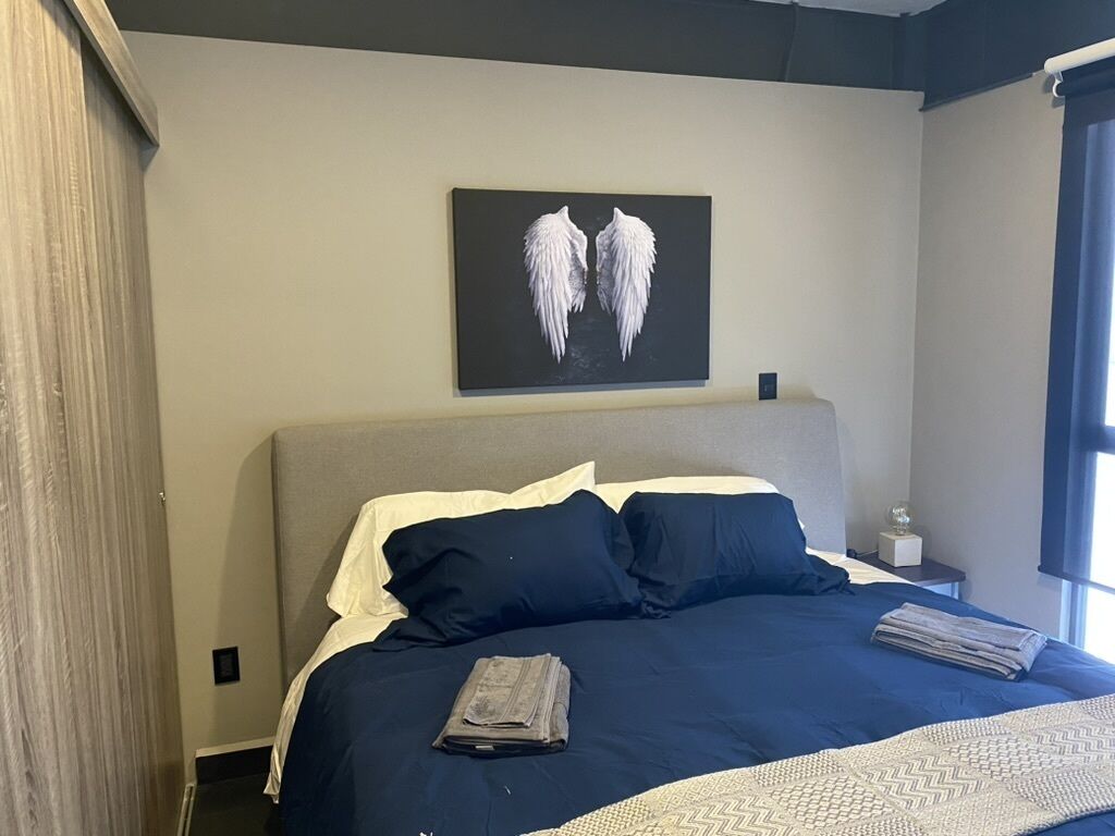 A neatly made bed with a dark blue bedspread and white pillows, a wall-mounted artwork of white wings, and a small bedside table with a glass.