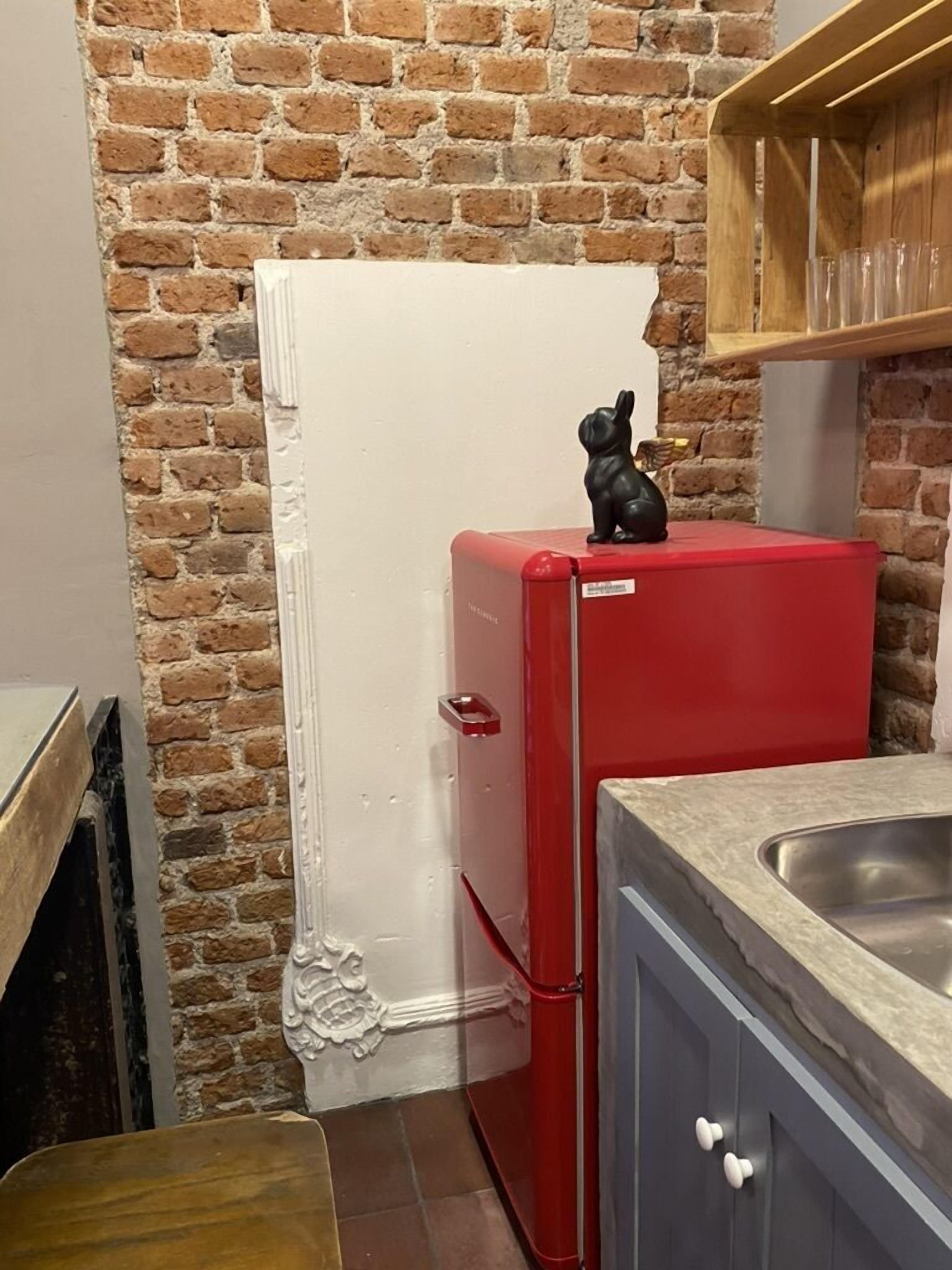 A kitchen with a brick wall, a red refrigerator, a sink, and a wooden shelf.
