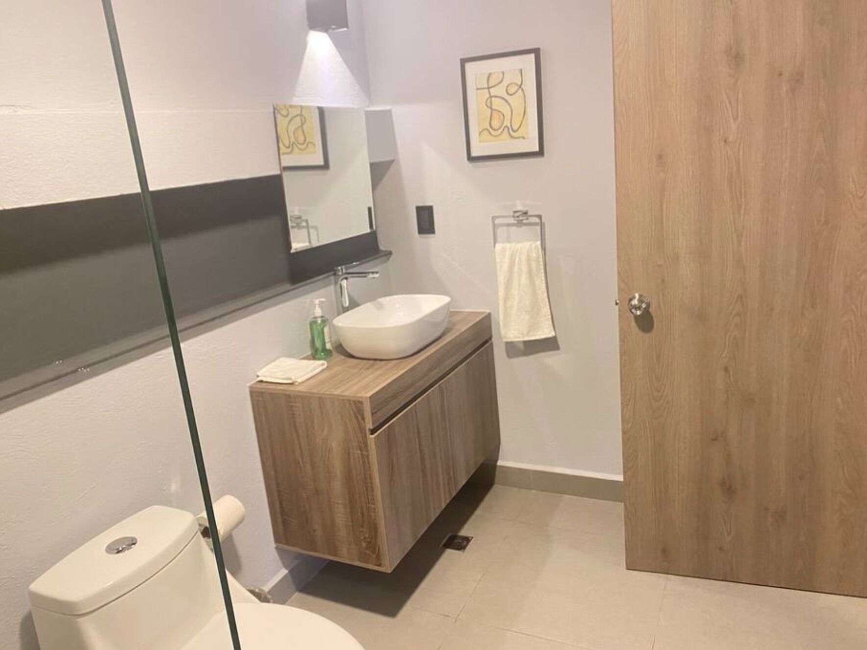 Un baño moderno con inodoro blanco, tocador de madera y lavamanos blanco, y cabina de ducha con paredes de vidrio.