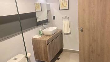 Un baño moderno con inodoro blanco, tocador de madera y lavamanos blanco, y cabina de ducha con paredes de vidrio.