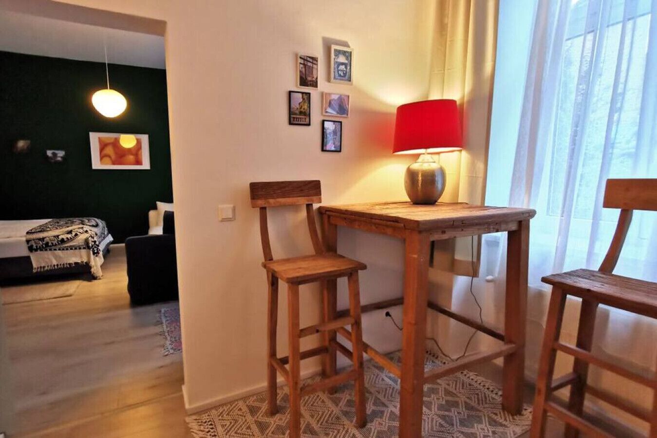 Une pièce avec une table en bois et deux chaises, une lampe rouge et un lit recouvert d’une couverture à motifs.