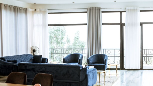 Un salón moderno con un sofá azul oscuro, dos sillones, una mesa de comedor y un ventilador. Amplias ventanas con cortinas dejan entrar luz natural y ofrecen vistas al exterior.