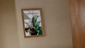 Una cama bien hecha con sábanas blancas, un cabecero de madera y una mesita de noche pequeña con un cajón. Sobre la cama, en la pared, cuelga un cuadro con plantas verdes.