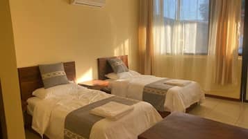 A hotel room with two beds, a wooden headboard, a bedside table, an air conditioning unit, and a large window with curtains.