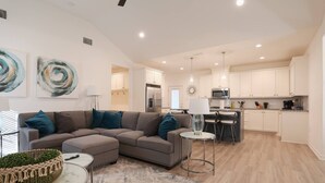 Ein modernes Wohnzimmer mit einem grauen Ecksofa, einem kleinen runden Tisch und einem Couchtisch. Im Hintergrund befindet sich eine Küche mit weißen Schränken und einem Essbereich.