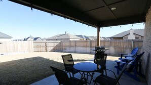 Una veranda coperta con tavolo e sedie, un barbecue e vista su un giardino sul retro con una recinzione in legno e case sullo sfondo.