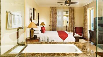 Habitación de hotel con un baño grande de mármol, una cama con ropa blanca y un plaid rojo, dos lámparas de noche y un ventilador de techo.