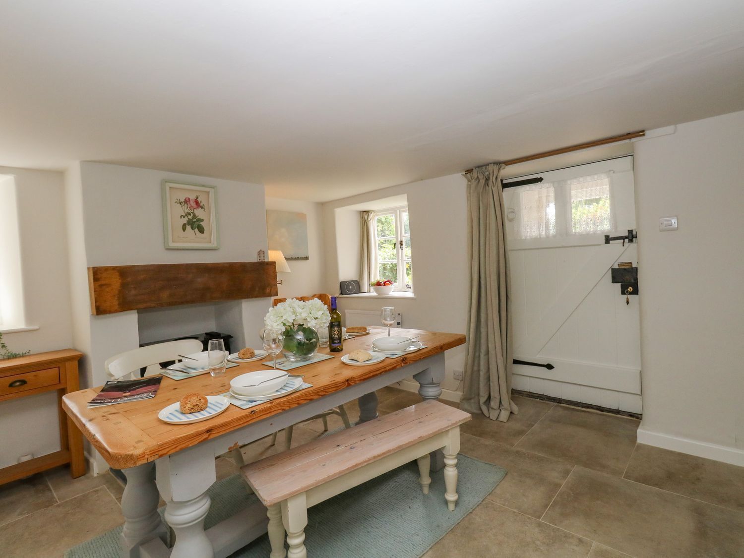 A dining area with a wooden table set for two, a bench, and a window with curtains.