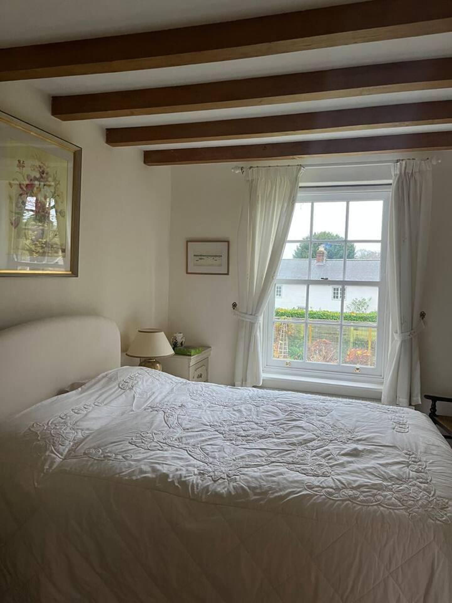 Ein Schlafzimmer mit einem großen Bett, einem Fenster mit Vorhängen, einem Nachttisch mit Lampe und einem gerahmten Bild an der Wand.
