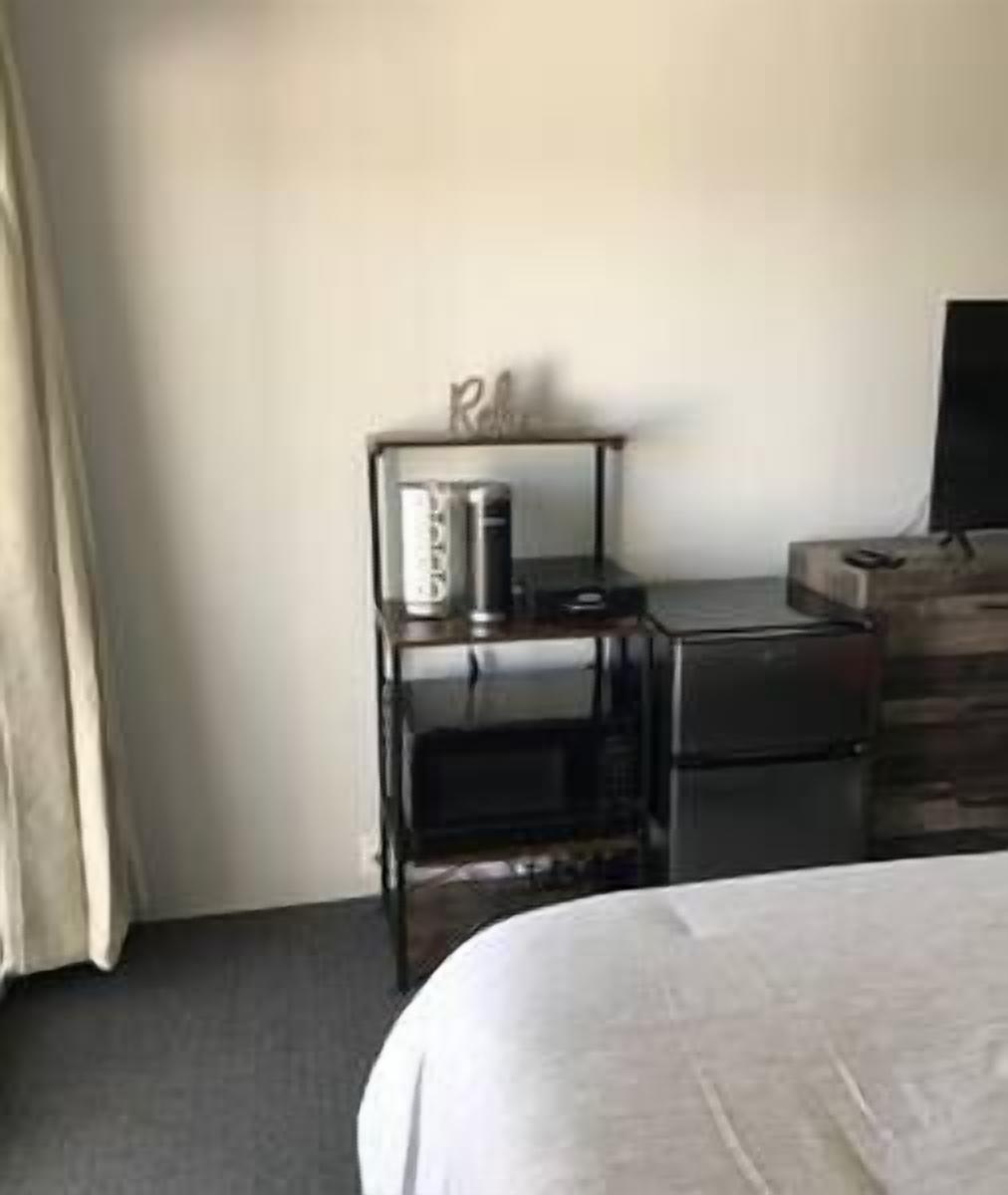 A hotel room with a bed, a desk with a computer and a printer, and a small shelf with a lamp and a decorative item.