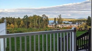 Näkymä parvekkeelta, jossa on valkoinen kaide. Parvekkeelta avautuu näkymä järvenrantapuistoon, jossa on puita ja kauempana näkyy rakennuksia.