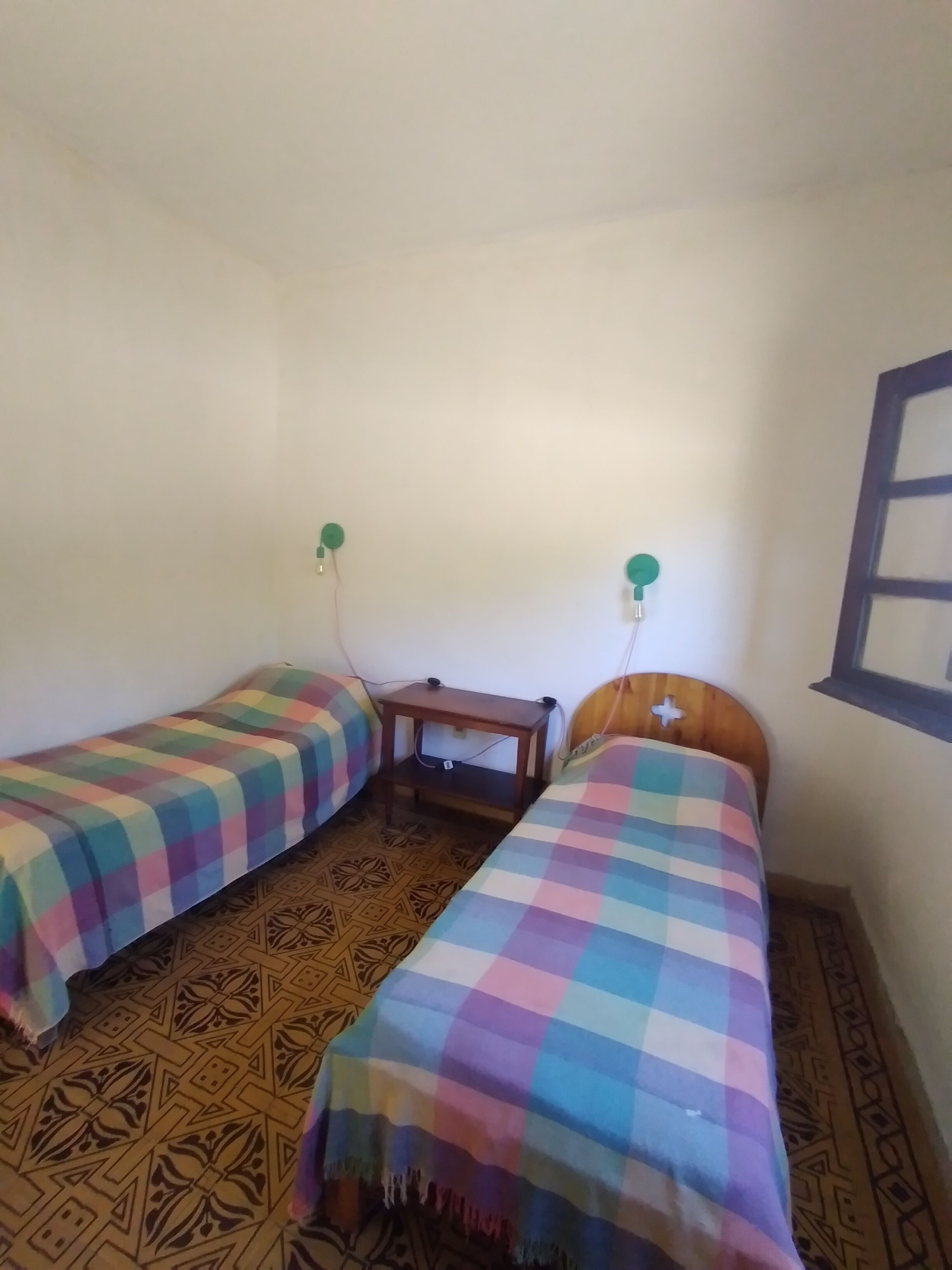 A small room with two single beds, each with a colorful checkered bedspread. There is a wooden nightstand between the beds with a lamp on it. The floor has a geometric patterned design.