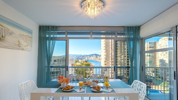 Un comedor con una mesa dispuesta para el desayuno, ventanas amplias con vistas a la ciudad y la playa, y una araña colgante.