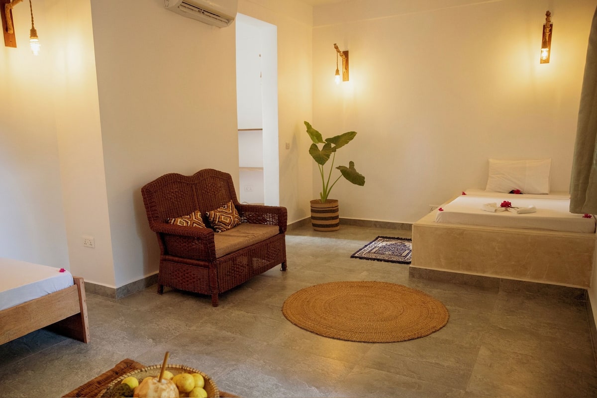 Ein Zimmer mit einem Korbstuhl, einem kleinen Bett, einer großen Topfpflanze und einem runden Teppich.