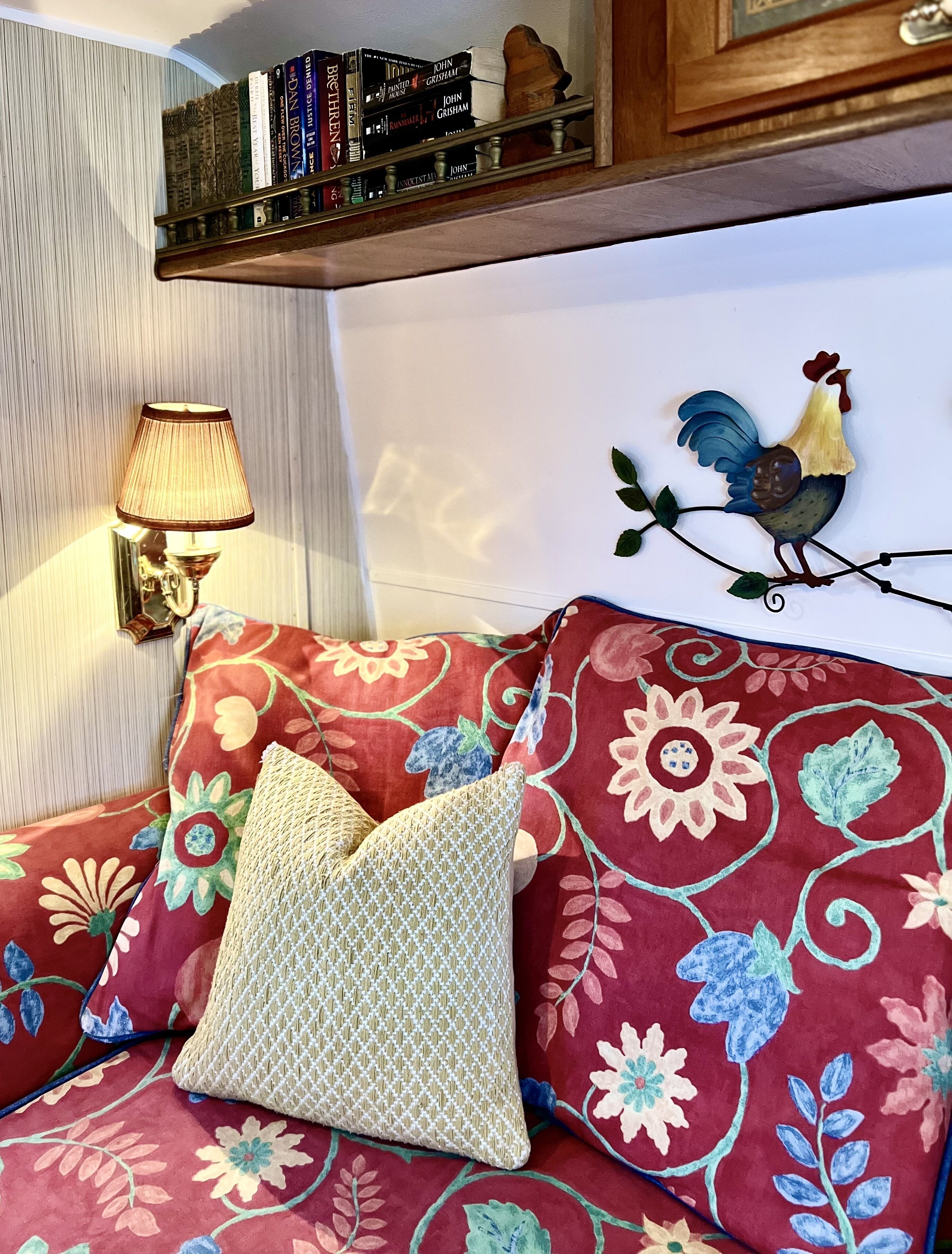 A room with a floral-patterned sofa, a decorative rooster wall art, a bookshelf with books, and a wall-mounted lamp.