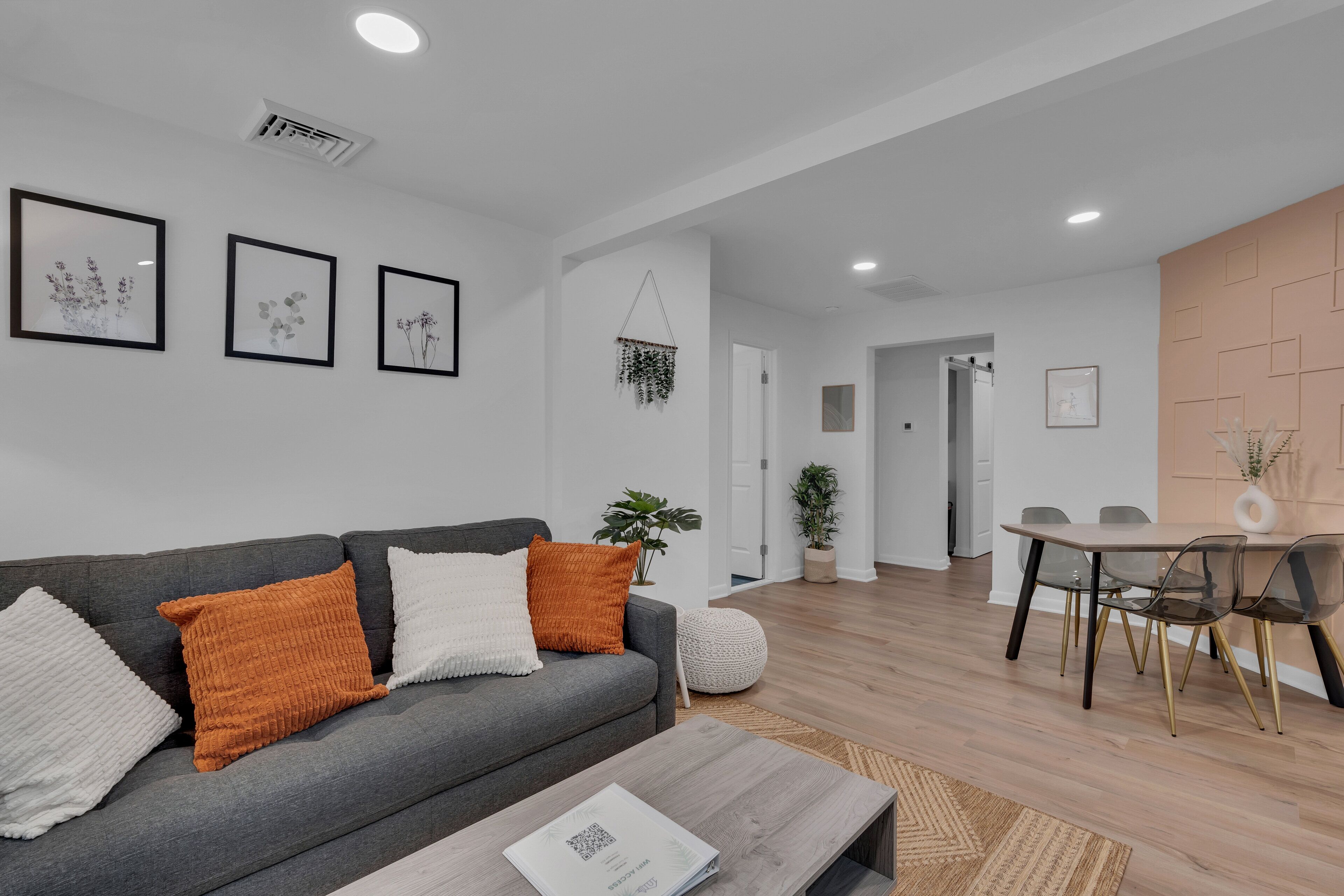Ein modernes Wohnzimmer mit grauem Sofa, weißen und orangefarbenen Kissen, einem Holztisch und einem Essbereich mit Holztisch und Stühlen.