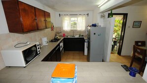A compact kitchen with a microwave, sink, and refrigerator. There is a window with curtains, a wooden cabinet, and a small dining area visible.
