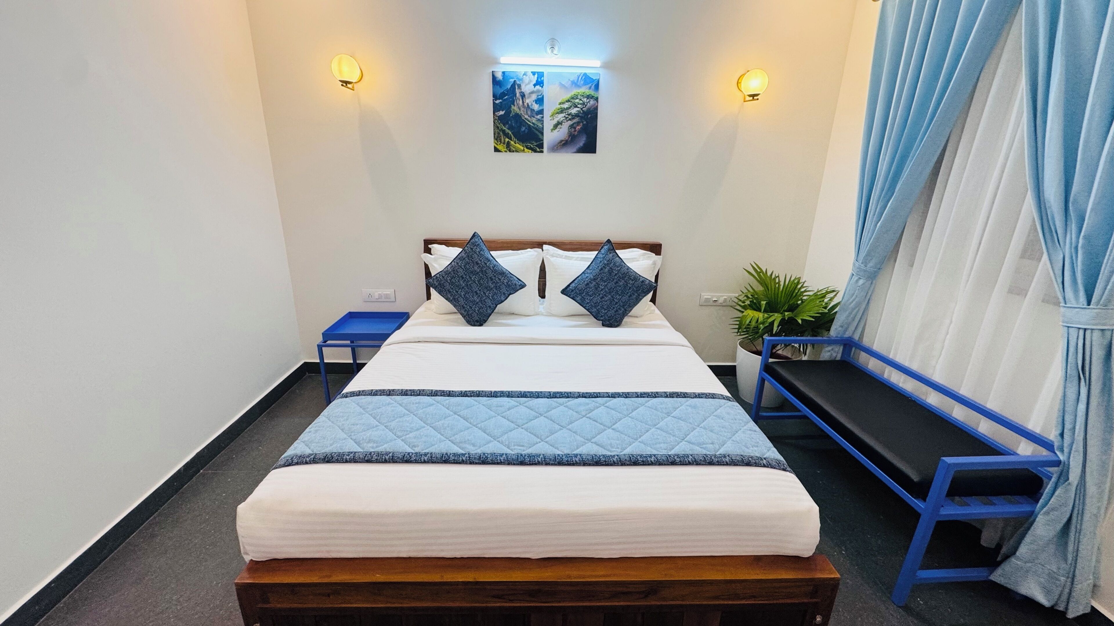 A hotel room with a bed, two wall-mounted lights, a small table, a bench, and a potted plant.