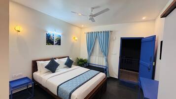 Um quarto com cama, ventilador de teto, janela com cortinas, porta e uma pequena mesa.