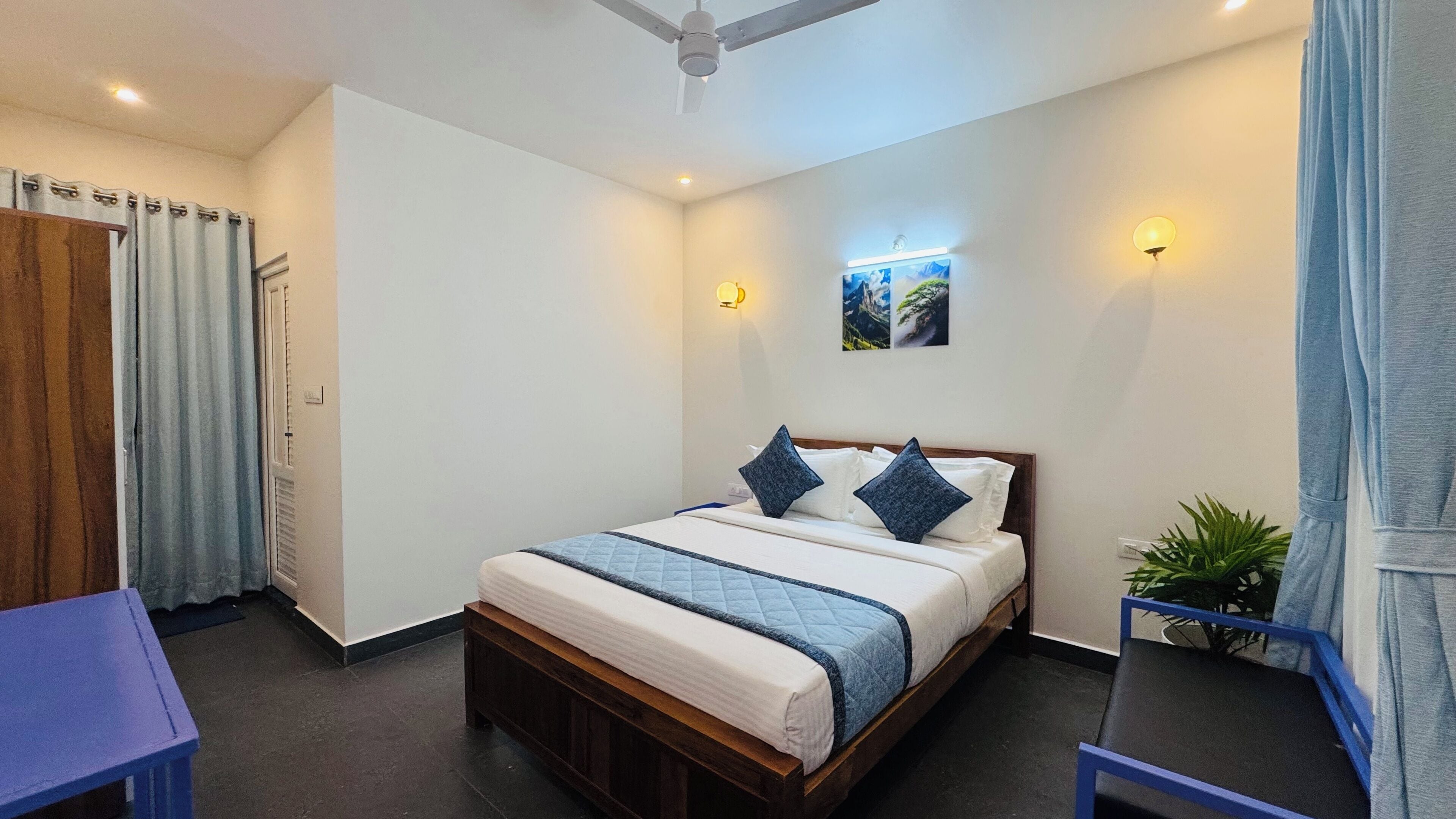 A bedroom with a bed, a ceiling fan, a wall-mounted light, a small potted plant, and a window with curtains.