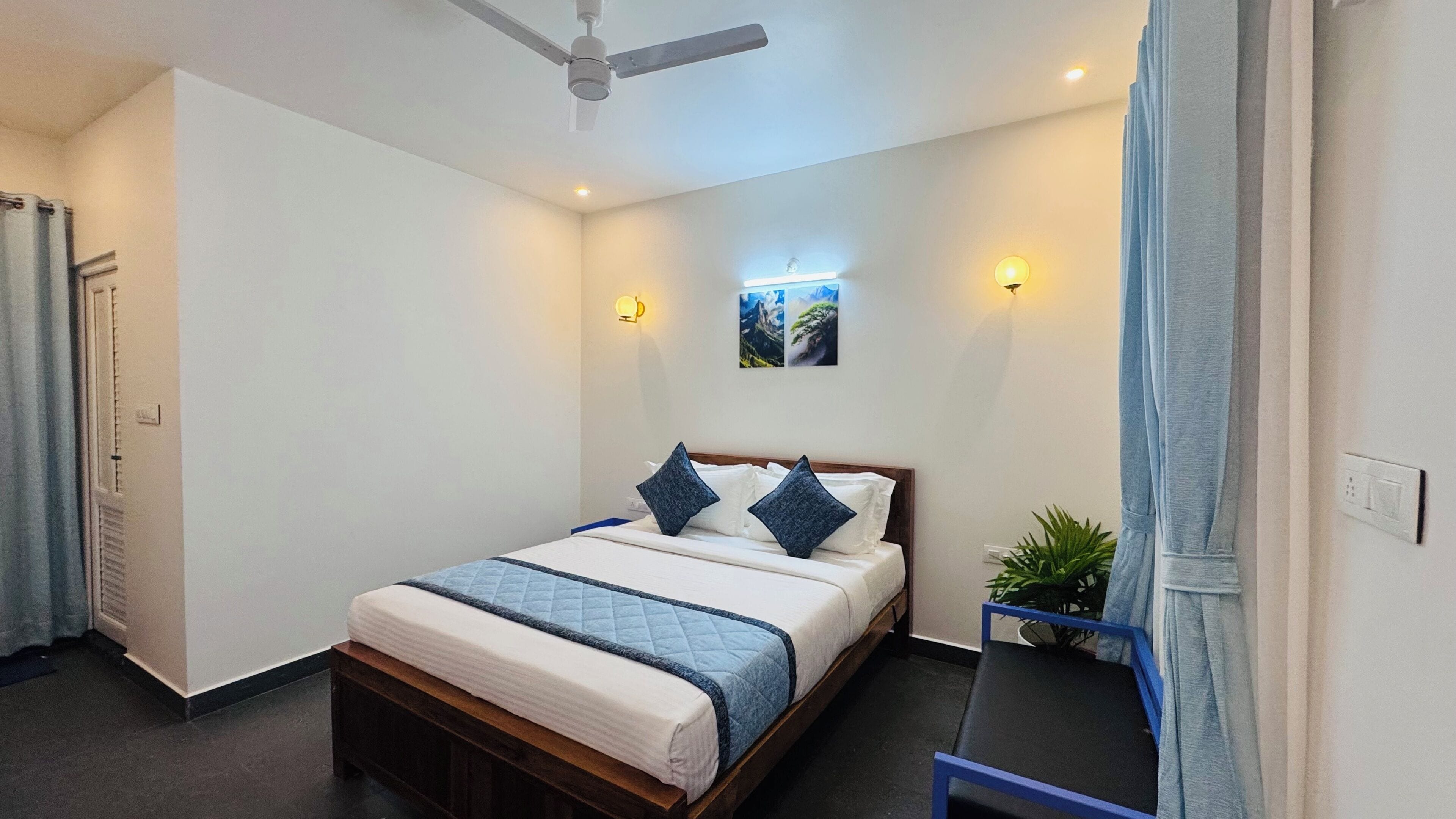 Una cama bien hecha con cubrecama acolchada y dos almohadas, un ventilador de techo, una mesita de noche con una planta, y una lámpara de pared.