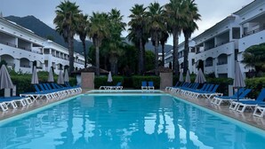A swimming pool with lounge chairs and umbrellas, surrounded by palm trees and a building with multiple balconies. - Appartement VUE MER Dans Résidence Standing Avec Piscine (San-Nicolao)