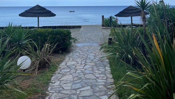 A stone pathway leading to a beach with two thatched umbrellas and lush greenery. - Appartement VUE MER Dans Résidence Standing Avec Piscine (San-Nicolao)