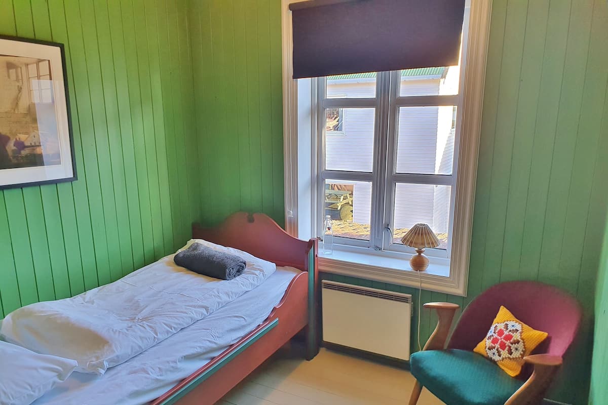 A small, green-walled room with a single bed, a chair, and a window with a view of a house.