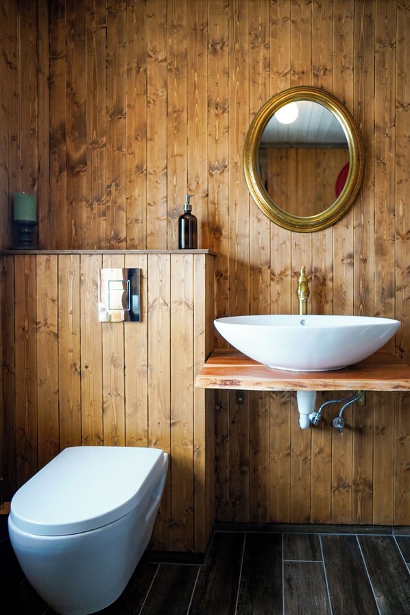 Un baño con paneles de madera, un inodoro blanco, un lavabo blanco con grifo dorado y un espejo redondo.