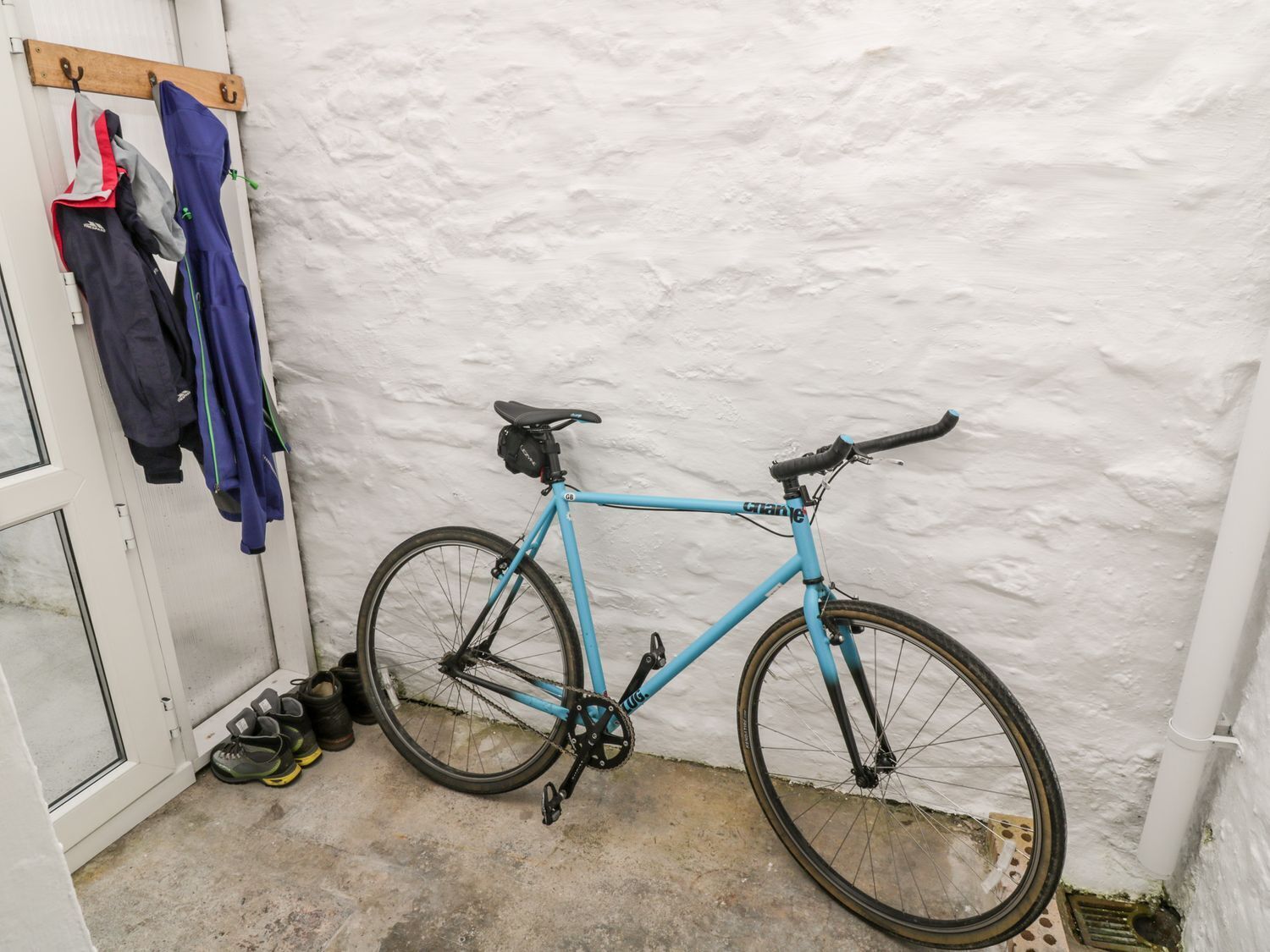 Een blauwe fiets staat tegen een witte muur in een hoek, met een schoenenrek met verschillende schoenen en jassen.