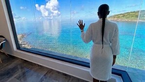 A person in a white robe standing by a large window overlooking a body of water and a distant landmass. - Condostmaarten panoramic appartment (Adults Only) (Sint Maarten)