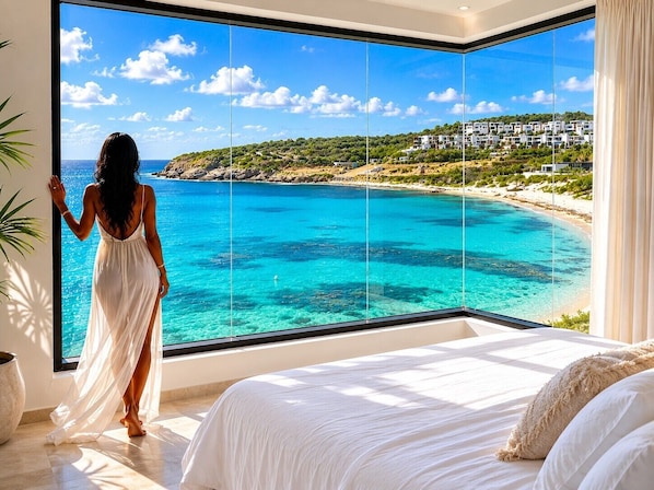 A bedroom with a large window overlooking a beach and a residential area.
