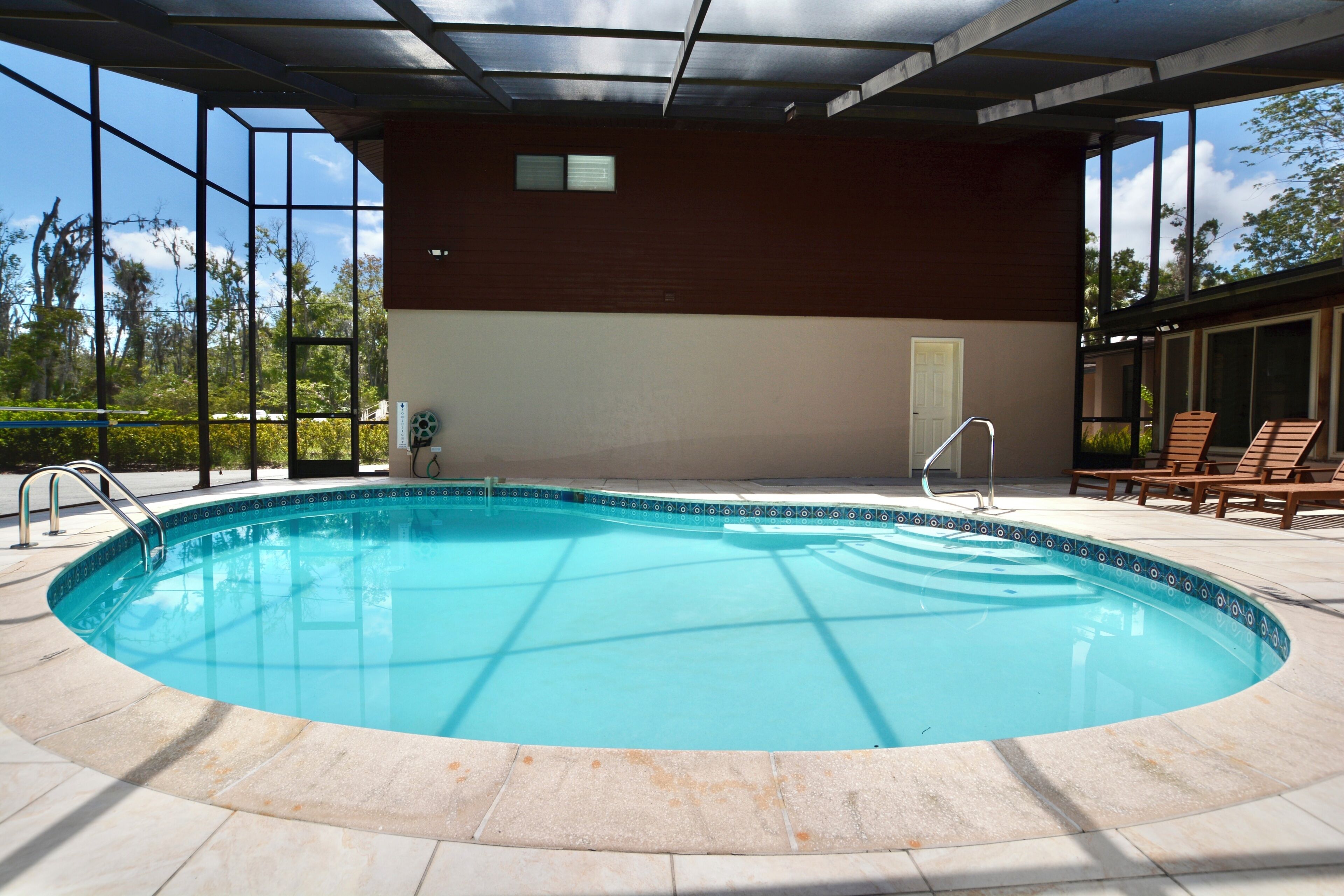 A swimming pool with a tiled deck, a covered area with a door, and a view of trees through large windows.