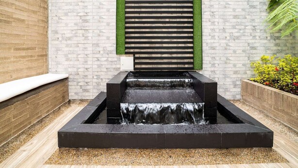 Una moderna fuente exterior con diseño escalonado, frente a un muro de ladrillo con un elemento vertical verde.