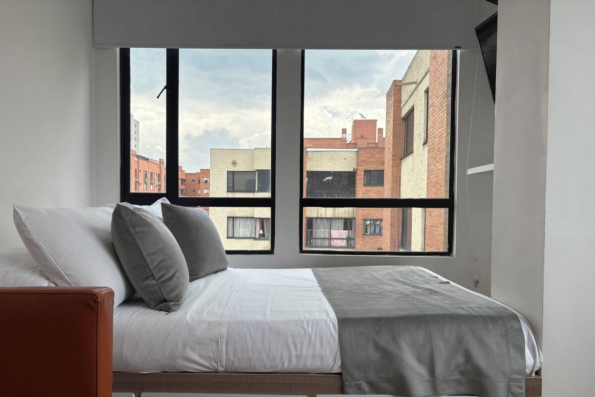 Een slaapkamer met een groot raam, een bed met wit en grijs beddengoed, en een uitzicht op gebouwen.