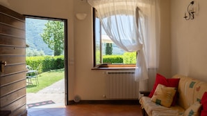Een kamer met een houten deur, een raam met lichte gordijnen, een radiator, een bank met kussens en uitzicht op een tuin en heuvels.