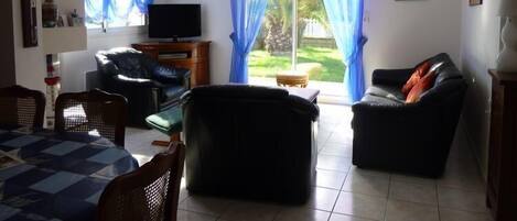 Un salón con televisor, dos sofás, una mesa de comedor y vistas a un jardín a través de amplias ventanas.