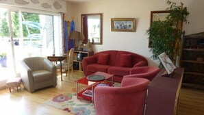 Un salón con un sofá rojo, un sillón beige, una mesita y una estantería. Se ve una ventana con vistas a zonas verdes.