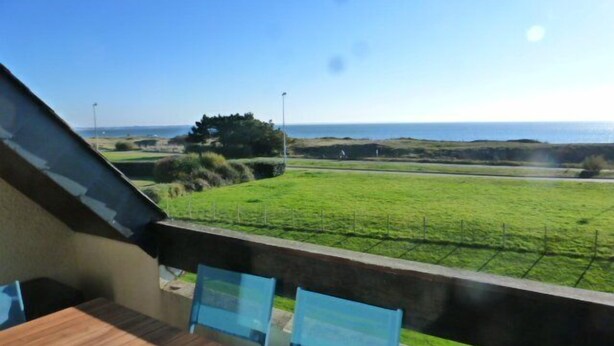 Ein Balkon mit einem Holztisch und zwei blauen Stühlen, mit Blick auf ein Wiesenfeld und das Meer.