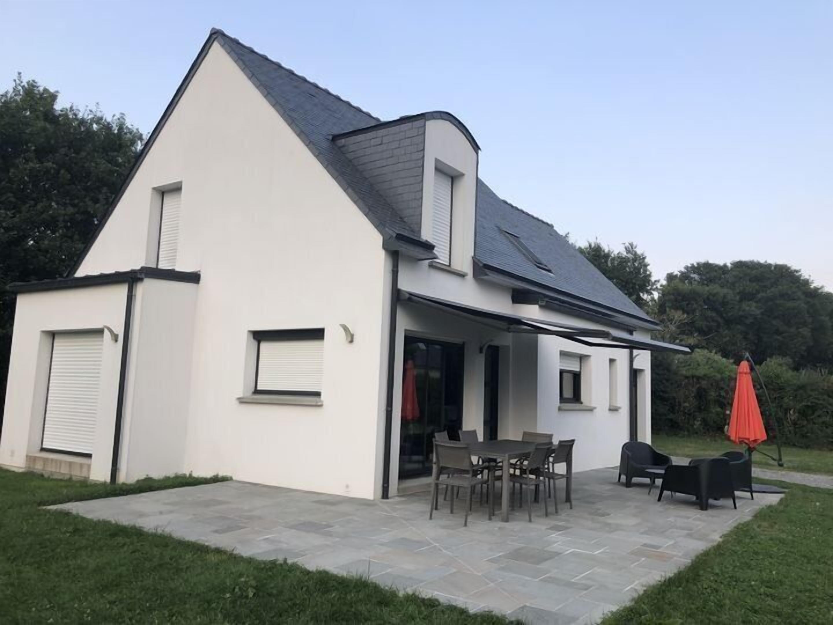 Ein modernes, zweistöckiges Haus mit einer Terrasse, die einen Tisch, Stühle, einen roten Sonnenschirm und eine Terrasse mit Steinplatten aufweist.