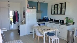 Ett kompakt kök med vita skåp, ett matbord med stolar, en hängande lampa och en papperskorg.